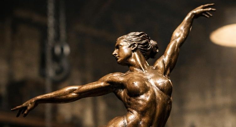 Sculpture figurative en bronze d'une femme nue en mouvement de danse classique, exposée dans un atelier d'Ayla Woss avec outils de forge en arrière-plan.
