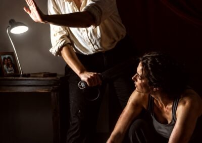 Photographie d'art dramatique représentant deux femmes dans une pièce sombre, l'une debout avec une main tendue pour guetter dans l'ombre tout en tenant une ceinture, l'autre accroupie rassemblant leurs affaires. Hommage à Artemisia Gentileschi.