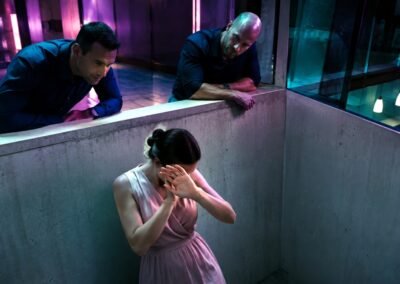 Photographie d'art montrant une femme en robe rose pastel acculée contre un mur en béton, se protégeant le visage de ses mains sous le regard dominant et manipulateur de deux hommes au-dessus d'elle.